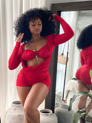 Woman wearing a bold red two-piece-style romper featuring a ruched, form-fitting design with long sleeves, a dramatic front cutout, and a central gold clasp detail. The outfit is stylish and modern, accentuating curves with gathered fabric and a vibrant color.