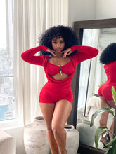 Woman wearing a bold red two-piece-style romper featuring a ruched, form-fitting design with long sleeves, a dramatic front cutout, and a central gold clasp detail. The outfit is stylish and modern, accentuating curves with gathered fabric and a vibrant color.