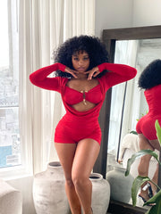 Woman wearing a bold red two-piece-style romper featuring a ruched, form-fitting design with long sleeves, a dramatic front cutout, and a central gold clasp detail. The outfit is stylish and modern, accentuating curves with gathered fabric and a vibrant color.