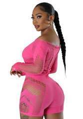 Woman wearing a hot pink mesh cutout romper with long sleeves and off-the-shoulder neckline, featuring intricate knit patterns and body-hugging fit, accessorized with gold heels and a metallic handbag.