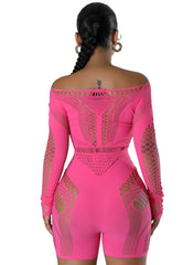 Woman wearing a hot pink mesh cutout romper with long sleeves and off-the-shoulder neckline, featuring intricate knit patterns and body-hugging fit, accessorized with gold heels and a metallic handbag.