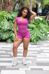A woman posing outdoors in a bright pink two-piece athletic set consisting of a scoop-neck short-sleeve top and matching high-waisted shorts, paired with white socks and sneakers, set against a background of green tropical plants and a striped pavement.