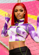Woman wearing a long sleeve purple and white tie-dye mock neck crop top with neon green contrast stitching, standing in front of a colorful fence.
