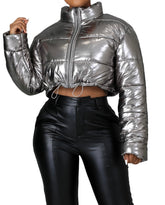 Woman wearing a metallic silver cropped puffer jacket with front zipper closure and drawstring hem, paired with high-waisted black faux leather pants.