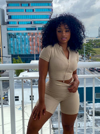 Woman in a beige ribbed two-piece set featuring a zip-up short-sleeve crop top and high-waisted biker shorts.