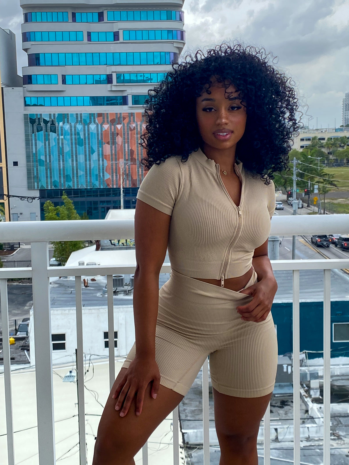 Woman in a beige ribbed two-piece set featuring a zip-up short-sleeve crop top and high-waisted biker shorts.