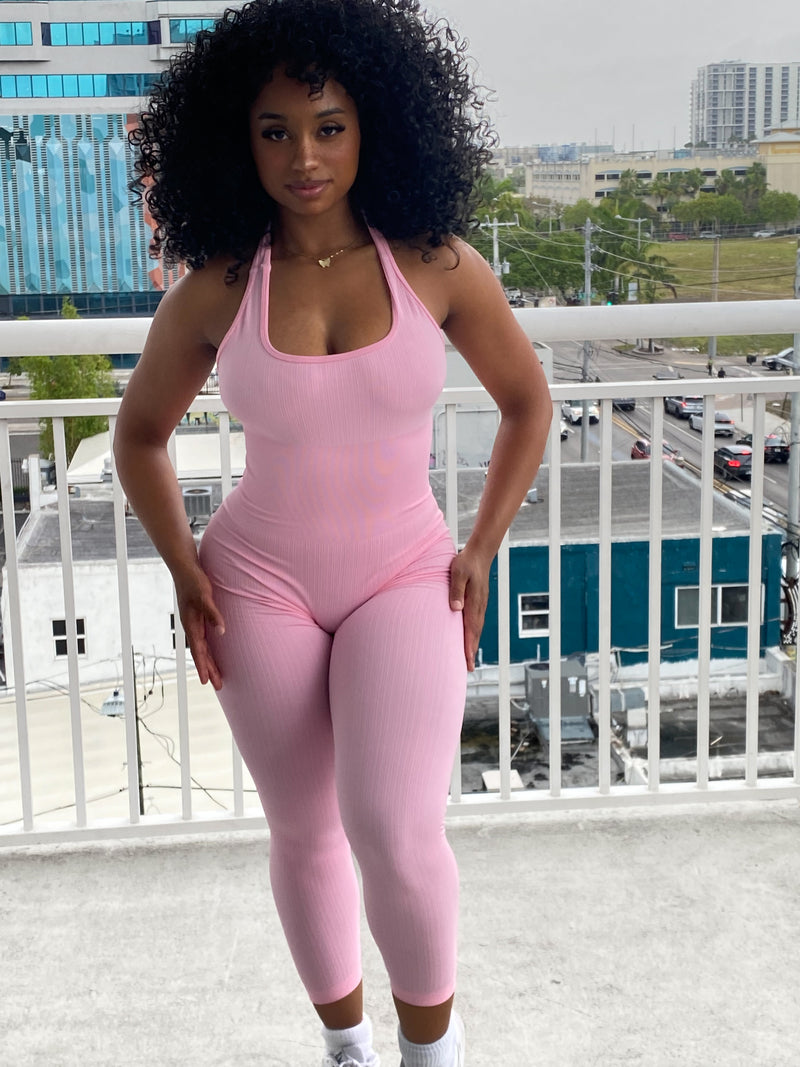 Woman wearing a fitted light pink halter jumpsuit with ribbed fabric, ankle-length leggings, and a scoop neckline.