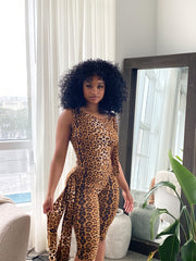 Woman wearing a sleeveless leopard print romper with side tie accents, fitted biker short length, posing indoors in front of a mirror with decorative plants and vases in the background.
