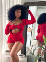 Woman wearing a bold red two-piece-style romper featuring a ruched, form-fitting design with long sleeves, a dramatic front cutout, and a central gold clasp detail. The outfit is stylish and modern, accentuating curves with gathered fabric and a vibrant color.