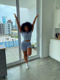 Woman wearing a baby blue ribbed two-piece set with a short-sleeve zip-up crop top and matching high-waisted biker shorts.
