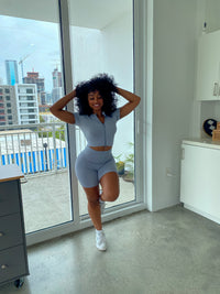 Woman wearing a baby blue ribbed two-piece set with a short-sleeve zip-up crop top and matching high-waisted biker shorts.