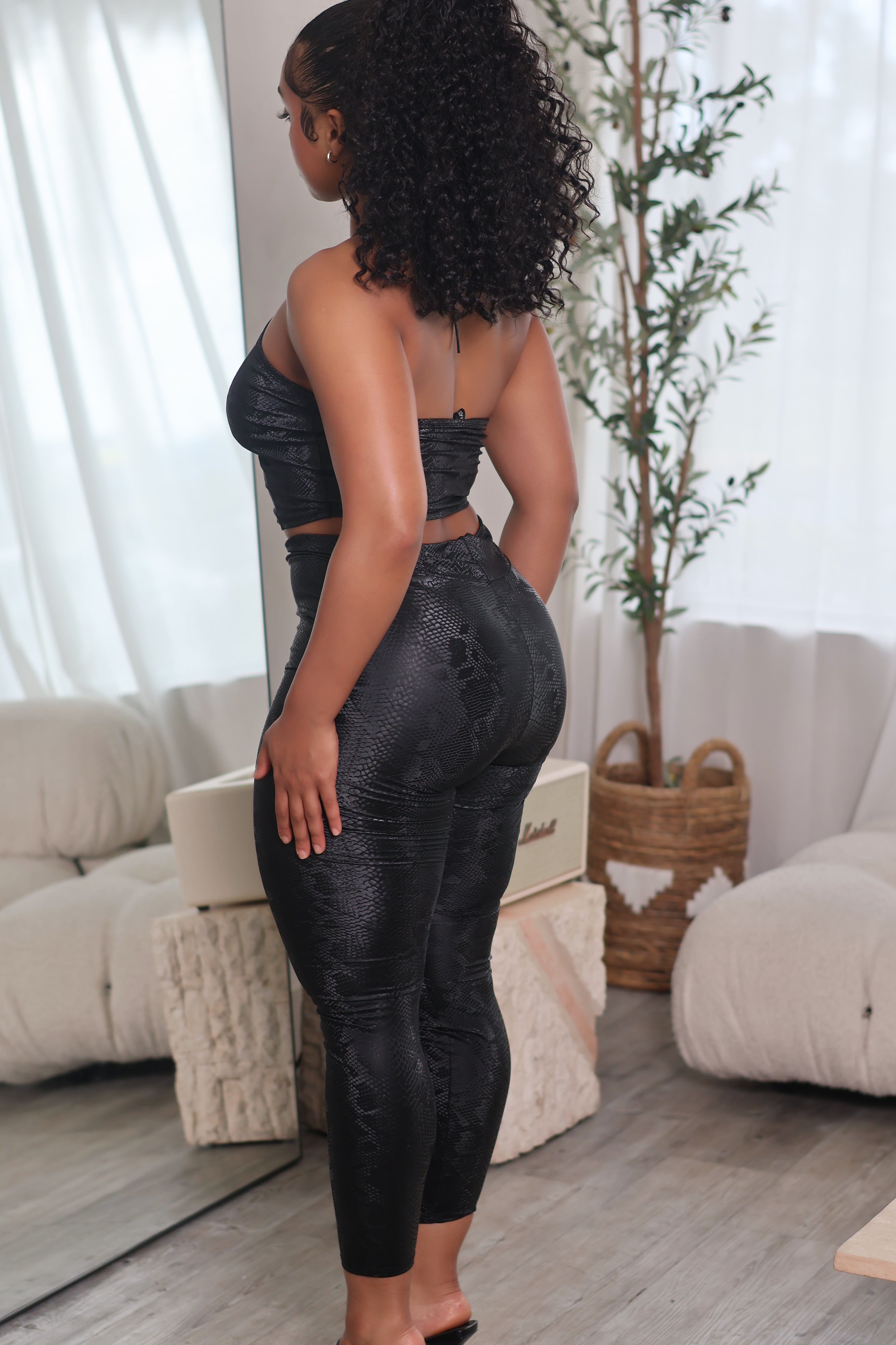 A woman stands in a bright room wearing a black textured two-piece outfit consisting of a strapless crop top and high-waisted leggings. She is shown from a rear three-quarter angle, with a mirror, beige sofas, and a potted plant in the background.