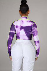 Woman wearing a long sleeve purple and white tie-dye mock neck crop top with neon green contrast stitching, standing in front of a colorful fence.