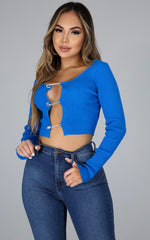 Woman wearing a long sleeve ribbed knit crop top with rhinestone safety-pin closures down the front, paired with high-rise skinny blue jeans.