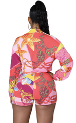 Woman wearing a colorful long-sleeve tie-front crop top and matching printed high-waist shorts with a bold baroque and chain pattern.