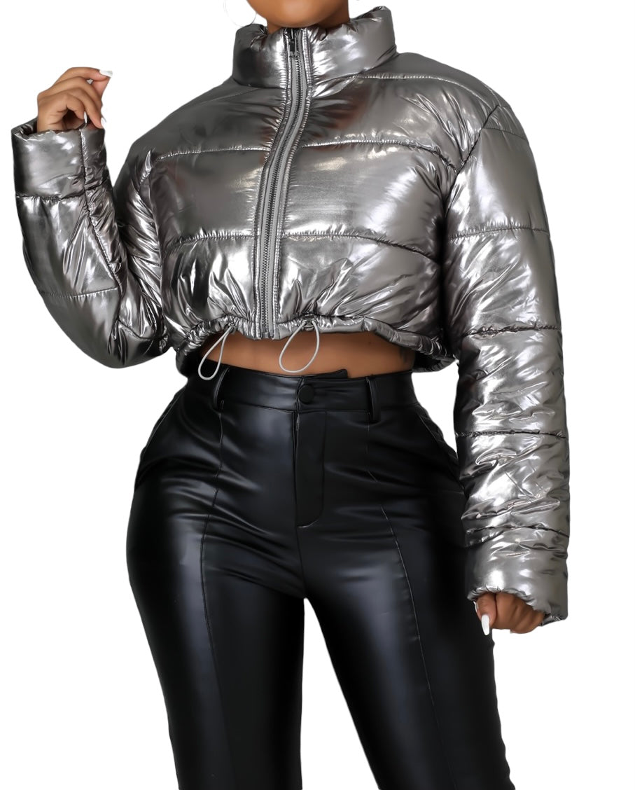 Woman wearing a metallic silver cropped puffer jacket with front zipper closure and drawstring hem, paired with high-waisted black faux leather pants.
